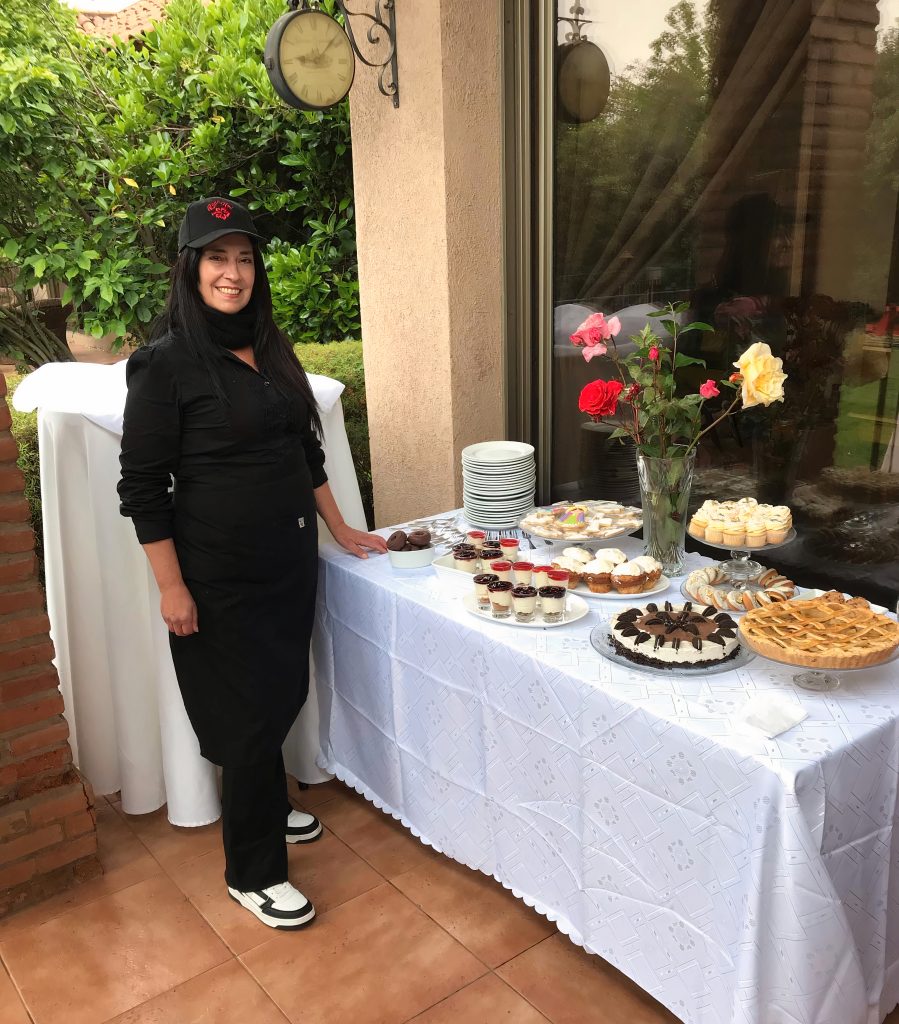 Carolina Guerrero vestida con un atuendo negro de catering junto a una mesa de postres cubierta con un mantel blanco, coronada con varios postres, pasteles y flores, situada al aire libre junto a un edificio con un jardín al fondo.